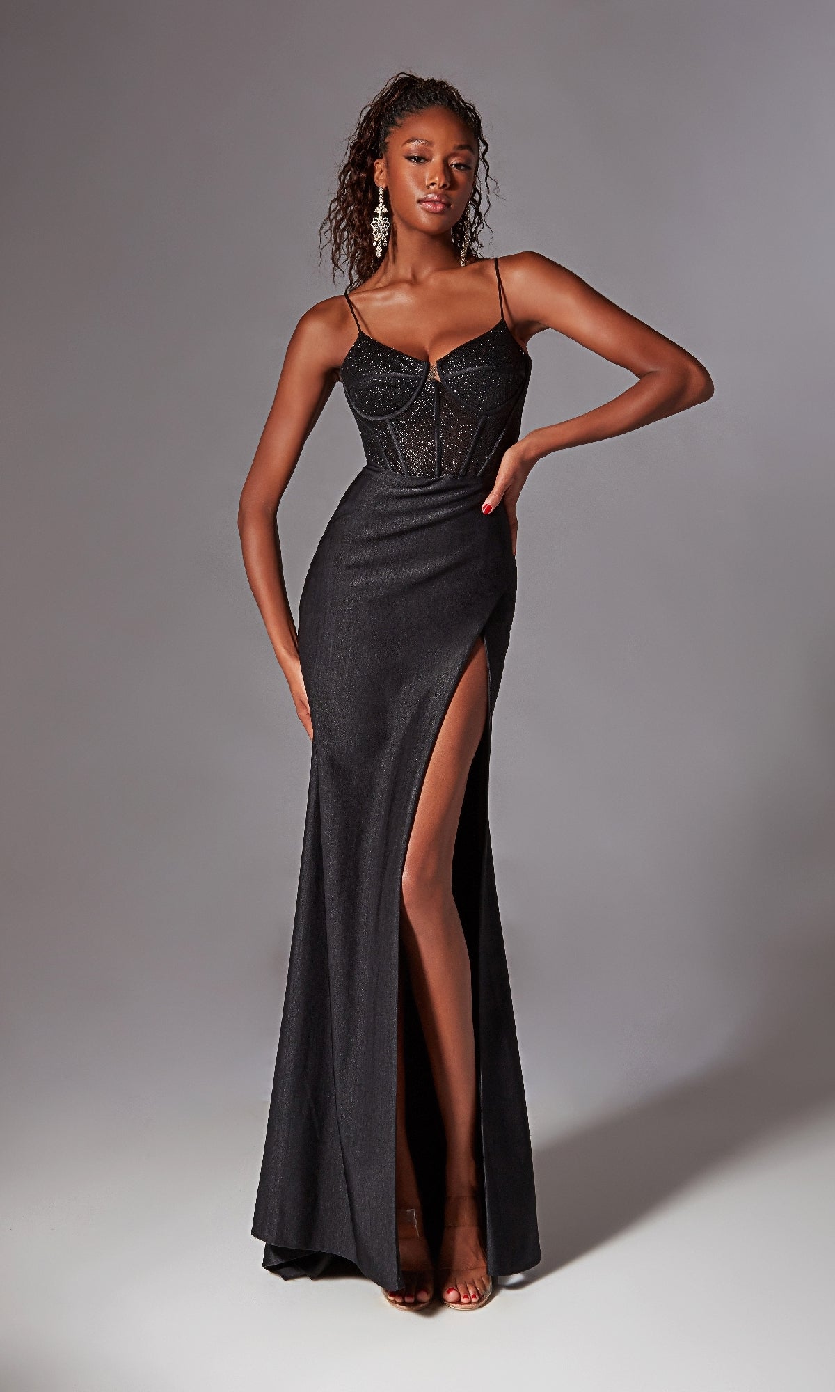 A woman models the Aerie Couture 269 Corset-Bodice Long Prom Dress—black, floor-length with glitter, spaghetti straps, sheer corset-style bodice, and a thigh-high slit—posing against a plain gray background.